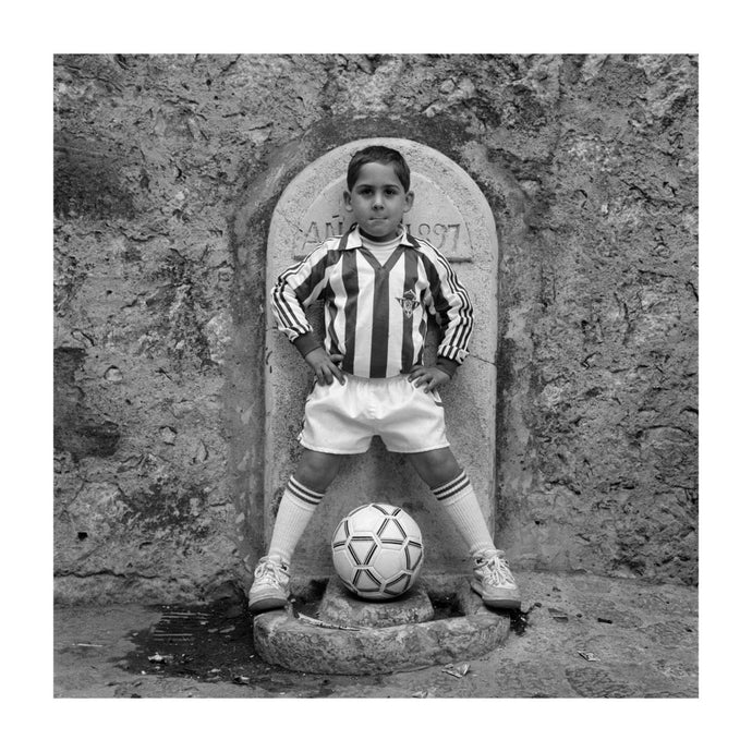 Black and white portrait of a young boy in Real Betis Balompie football kit, standing in front of a fountain. Pollensa,  Majorca 1992.